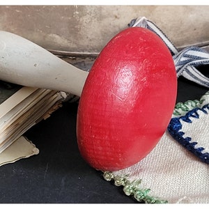 May include: A red wooden egg-shaped toy with a white wooden handle. The toy is sitting on a white cloth with green and blue trim.