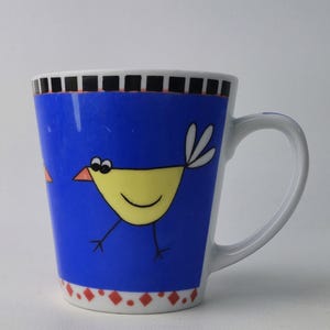 Peut inclure: Une tasse en céramique blanche avec un fond bleu représentant un oiseau de dessin animé jaune avec un visage souriant. La tasse a une anse blanche et un bord décoratif avec des carrés noirs et des diamants rouges.