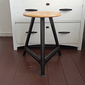 Op de afbeelding: Een zwarte metalen kruk met een houten zitting. De kruk heeft een driehoekige basis en staat op een donkerbruine houten vloer.