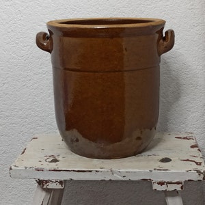 May include: A large, brown ceramic pot with two handles. The pot is sitting on a white, distressed wooden stool.
