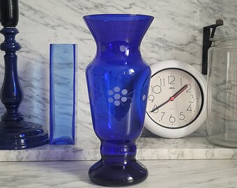 May include: A cobalt blue glass vase with a white floral design. The vase has a wide mouth and a round base.