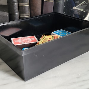 Può includere: Una scatola rettangolare nera con un coperchio, contenente diverse scatole di fiammiferi. Le scatole di fiammiferi hanno etichette colorate con testo, tra cui "Three Docks" e "Europa".