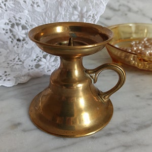 May include: A gold metal candle holder with a round base and a handle. The holder has a smooth, polished finish.