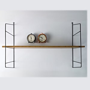 May include: Two alarm clocks, one black and one orange, sit on a wooden shelf with black metal brackets. The shelf is mounted on a white wall.