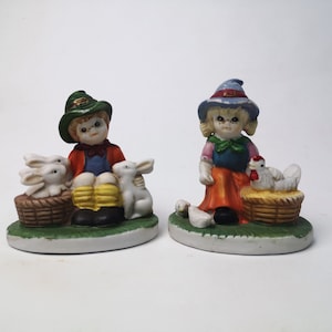 May include: Two ceramic figurines of children, one with a green hat and holding three white rabbits in a basket, the other with a blue hat and holding a white chicken in a basket.