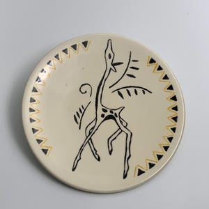 Op de afbeelding: Crèmekleurige keramische plaat met een zwart-geel geometrisch ontwerp. De plaat heeft een gestileerde zwarte tekening van een dier. De rand is versierd met afwisselende zwarte en gele driehoeken.