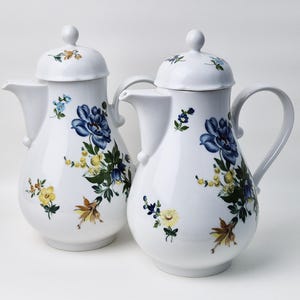 Könnte beinhalten: Zwei weiße Kaffeekannen aus Keramik mit Blumendesigns. Die Kannen haben blaue, gelbe und grüne Blumen auf weißem Hintergrund.
