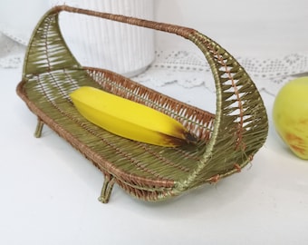 May include: A woven wicker fruit basket with a green and brown finish. The basket has a curved handle and stands on three legs. A yellow banana is resting inside the basket.