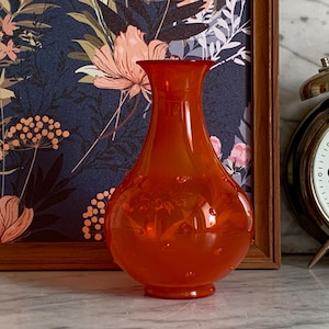Könnte beinhalten: Eine orangefarbene Glasvase mit bauchigem Körper und ausgestelltem Hals. Die Vase hat kleine, runde Blasen im Glas. Ein gerahmter Blumendruck und ein goldfarbener Wecker befinden sich im Hintergrund.