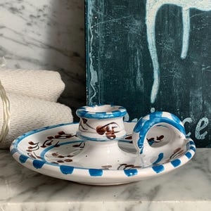 May include: A white ceramic candle holder with blue and brown accents. The holder has a circular base with a handle and a central cup for the candle. The design includes blue stripes and brown floral patterns. The background includes a marble surface and a teal painted board.