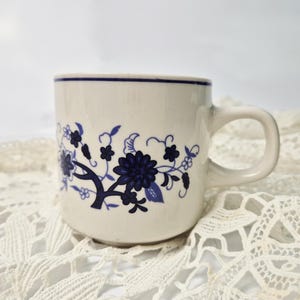 Può includere: Una tazza in ceramica bianca con un motivo floreale blu. La tazza ha un manico arrotondato e un bordo largo.