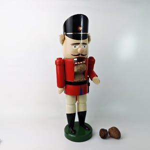 May include: A wooden nutcracker figurine wearing a red and black uniform, with a white face and a black hat. The nutcracker is holding a small object in its right hand. The nutcracker is standing on a green base.