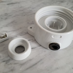 Może przedstawiać: Biała ceramiczna osłona gniazdka elektrycznego z okrągłym otworem i czarną gumową uszczelką. Osłona ma wypukły napis "M001".