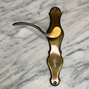 May include: A gold-toned door handle with a keyhole and a decorative design. The handle has a curved lever and a decorative plate.