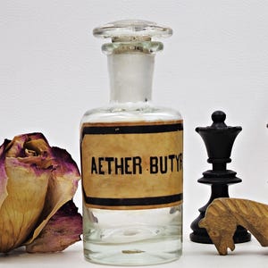 May include: A clear glass bottle with a cork stopper and a label that reads "AETHER BUTYR". The bottle is sitting on a white surface with a dried rose and chess pieces.