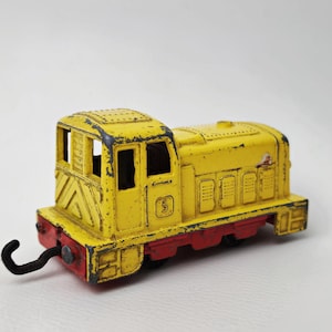 Könnte beinhalten: Ein gelber Spielzeug-Locomotive mit roten Rädern und einem schwarzen Haken. Die Lokomotive hat ein schwarz-weiß gestreiftes Muster an der Seite und eine Nummer "5" vorne.