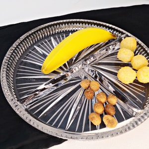 Op de afbeelding: Een glazen serveerschaal met drie vakken. De schaal heeft een decoratief stervormig patroon. Een gele banaan bevindt zich in het ene vak, kleine bruine snacks in het andere en kleine, ronde, gele snacks in het derde vak.