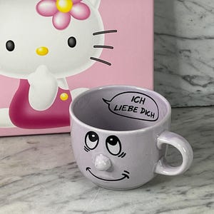 Könnte beinhalten: Ein hellvioletter Keramikbecher mit einer erhabenen Nase, einem aufgedruckten lächelnden Gesicht mit nach oben gerichteten Augen und dem Text "ICH LIEBE DICH" in einer Sprechblase am inneren Rand. Der Becher steht vor einer rosa Schachtel mit Hello Kitty.
