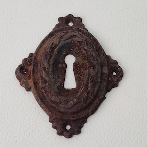 Può includere: Una placca per buco della serratura antica e arrugginita, con un centro ovale e bordi decorativi. Il metallo ha una superficie strutturata e un colore marrone scuro, con un'apertura a forma di buco della serratura al centro. Ha quattro fori di montaggio.