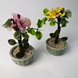 May include: Two small potted plants with green leaves and artificial flowers. One plant has pink flowers and the other has yellow flowers. The pots are green and have a textured surface.