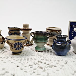May include: A collection of eight miniature ceramic pots in various styles and colors, including blue, green, brown, and white. The pots are decorated with floral and geometric patterns. The pots are arranged on a white crocheted surface.