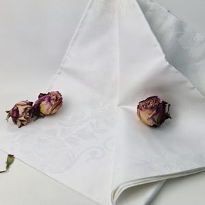 Könnte beinhalten: Gefaltete weiße Stoffserviette mit floralem Muster, verziert mit drei getrockneten Rosen. Die Rosen sind eine Mischung aus Lila, Gelb und Braun. Die Serviette ist dreieckig gefaltet.