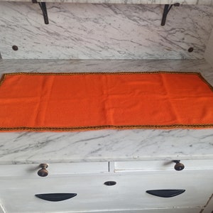 Könnte beinhalten: Orangefarbener gestrickter Platzdeckchen mit braunem und goldenem Rand. Das Platzdeckchen liegt auf einer weißen Marmorfläche.