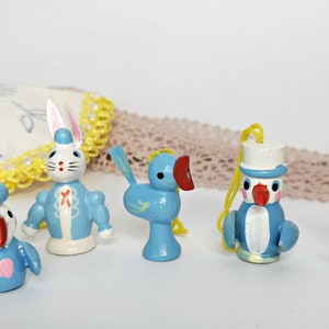 May include: Five wooden ornaments in blue, white, and red colors. The ornaments are shaped like a rabbit, a bird, a penguin, and two rabbits. The ornaments are all wearing hats.