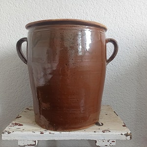 Può includere: Un grande vaso di ceramica smaltata marrone con due manici. Il vaso è appoggiato su una superficie di legno bianco invecchiato.