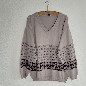 Könnte beinhalten: Ein hellbeiger Pullover mit V-Ausschnitt und einem braunen und weißen geometrischen Muster. Der Pullover hat lange Ärmel und einen gerippten Saum.