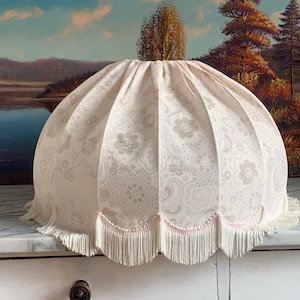 May include: A white lampshade with a scalloped edge and fringe trim. The lampshade has a floral pattern in shades of grey.