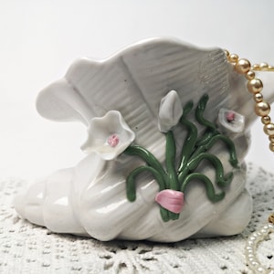 Peut inclure: Planteuse en céramique blanche en forme de coquille de mer avec des feuilles vertes et des fleurs roses et blanches. La planteuse est décorée d'un collier de perles.