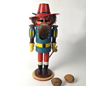 May include: A wooden nutcracker figurine with a red hat, blue uniform, and yellow accents. The nutcracker has a mustache and beard, and is holding a sword. There are three walnuts in the foreground.