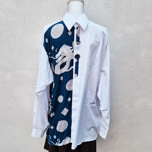 Könnte beinhalten: Weißes Button-Down-Hemd mit einem einzigartigen Design. Die linke Seite hat eine marineblaue Tafel mit einem weißen Drachen, Kreisen und geometrischen Formen. Das Hemd hat lange Ärmel und einen klassischen Kragen.