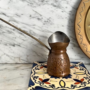 Könnte beinhalten: Eine türkische Kaffeekanne aus Kupfer mit einem langen Holzgriff. Der Topf hat florale Details und steht auf einer dekorativen Fliese. Im Hintergrund befindet sich eine Messingplatte mit einem geätzten Design.
