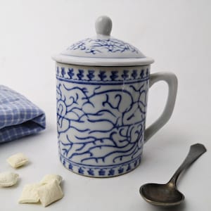 Op de afbeelding: Een witte keramische mok met een blauw en wit bloemenpatroon en een deksel. De mok heeft een handvat en staat op een wit oppervlak.