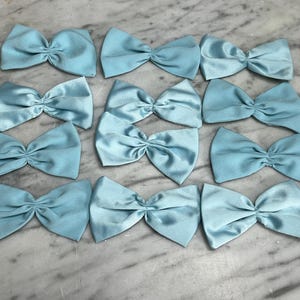 Op de afbeelding: Een verzameling van twaalf lichtblauwe satijnen strikken, elk in een klassieke strikvorm geknoopt. De strikken liggen op een marmeren oppervlak, wat hun delicate textuur en kleur benadrukt. Geschikt voor diverse knutselprojecten.