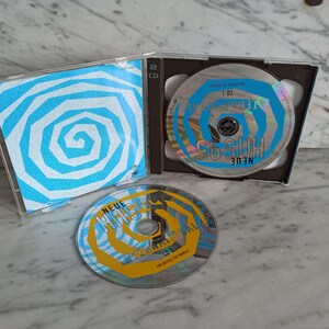 May include: Two CDs with blue and white spiral designs. The front of the CD case features a blue and white spiral design. The text on the CDs reads "Neue Hits 95" and "Neue Hits 95 - Die Ersten 100 Hits".