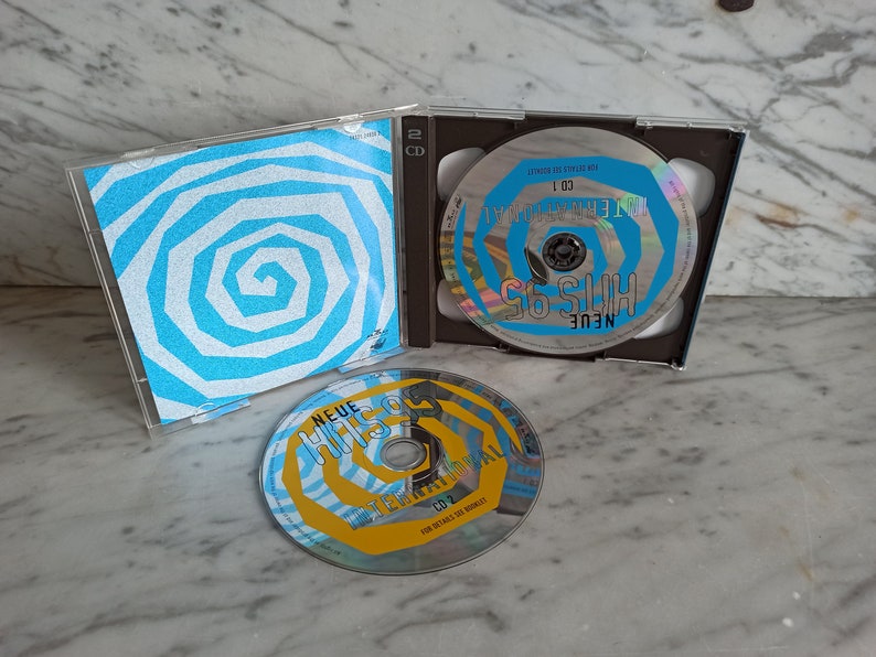 May include: Two CDs with blue and white spiral designs. The front of the CD case features a blue and white spiral design. The text on the CDs reads "Neue Hits 95" and "Neue Hits 95 - Die Ersten 100 Hits".
