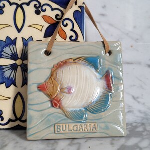 Può includere: Una piastrella di ceramica con un design di pesce blu, bianco e giallo. La piastrella è appesa a un cordino di cuoio marrone e ha la scritta "BULGARIA".