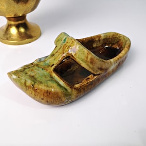 May include: A small, ceramic, green and brown shoe-shaped dish with a strap across the top. The dish has a glazed finish and is perfect for holding small items.