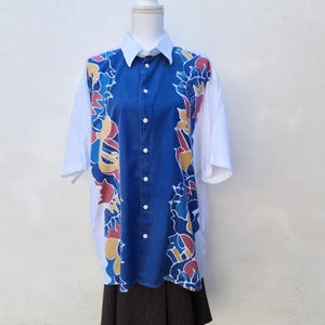 May include: A button-down shirt with a white collar and short sleeves. The front panel is a vibrant blue with a colourful floral design in red, yellow, and blue. The sides of the shirt are white.