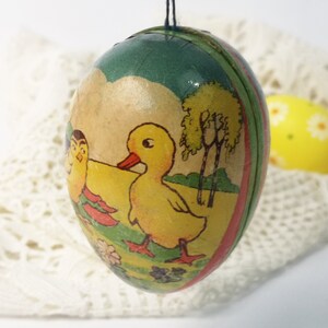 Può includere: Un uovo di Pasqua vintage in cartapesta con un'illustrazione colorata di un'anatra, un pulcino e alberi. L'uovo è decorato con vernice verde, gialla, rossa e bianca.