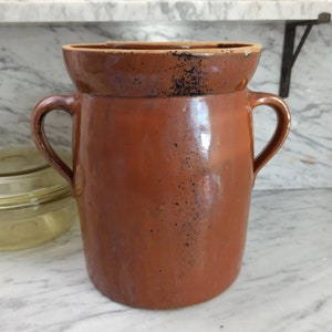 Può includere: Un grande vaso di terracotta marrone con due manici. Il vaso ha una finitura smaltata ed è leggermente macchiato di nero.