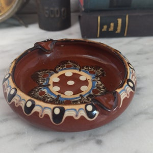 May include: A brown ceramic ashtray with a white and blue floral design and white polka dots. The ashtray has a rim with black and white accents.