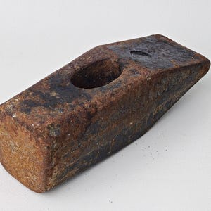 May include: A heavy-duty, rectangular sledgehammer with a rust-colored, weathered appearance. The hammerhead features a large, circular eye for the handle and is made of solid, dark metal. The tool is designed for demolition and heavy construction.