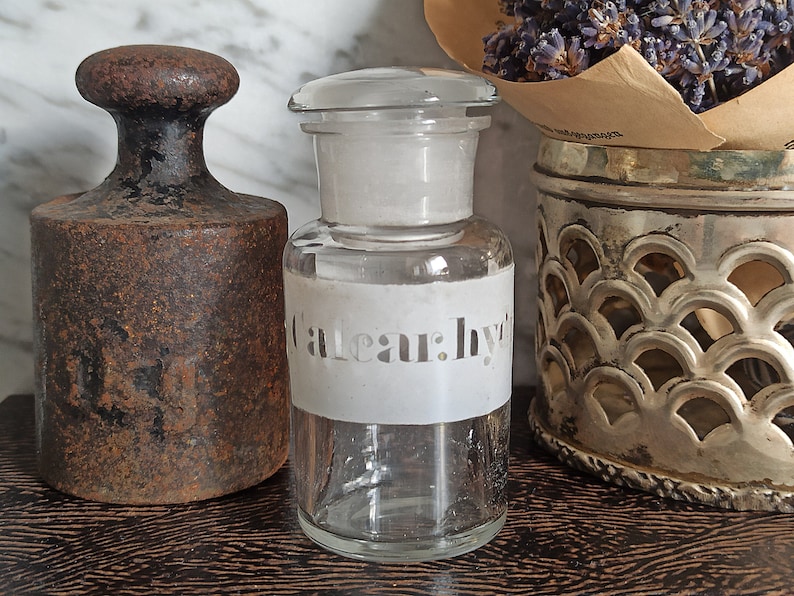 May include: A clear glass bottle with a white label that says "Eau: Hy" on it. The bottle is sitting on a wooden surface next to a rusty metal weight.