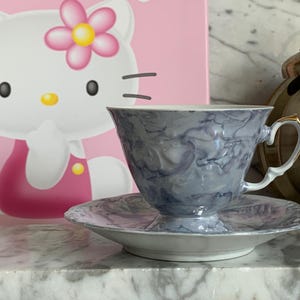 May include: A gray and white teacup and saucer with a marbled design. The teacup has a gold trim around the rim and a gold handle. The saucer has a matching marbled design.