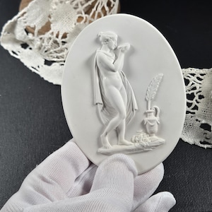 May include: A white oval plaque with a classical relief sculpture of a draped figure holding a vessel. A vase with a feather is also depicted. The plaque is held by a gloved hand, with lace in the background.