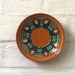 Peut inclure: Une assiette ronde en céramique avec un motif peint à la main. L'assiette présente un cercle orange central entouré d'un motif de formes bleues, vertes et blanches. Le bord est orange avec des accents blancs et bruns.